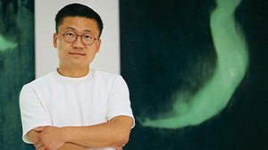 ie Lei, artiste contemporain, pose dans son atelier lumineux. En arrière-plan, deux grands tableaux sombres révèlent des silhouettes vertes spectrales, semblant flotter ou tomber, créant une ambiance onirique et mystérieuse.