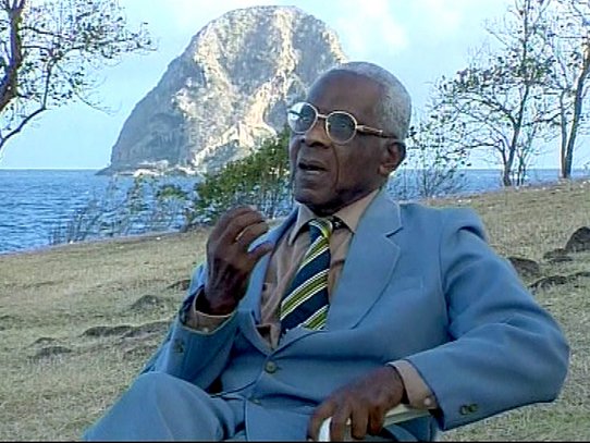 Sitting outdoors in a natural, dry landscape, an elderly man with short grey hair wears a neatly tailored light blue suit, a beige shirt, and a striped tie in shades of blue, green, and yellow. He speaks with emotion, his right hand raised in a thoughtful or expressive gesture, eyes directed toward the horizon. Behind him rises the iconic Diamond Rock, a volcanic islet off the coast of Martinique, standing firm in the turquoise sea. The arid ground, scattered with stones, contrasts with the vastness of the ocean, and a few sparse, leafless trees frame the scene. The moment captured exudes gravity and remembrance, reflecting the documentary’s mission to honor the memory of slavery and the voices that preserve it.
