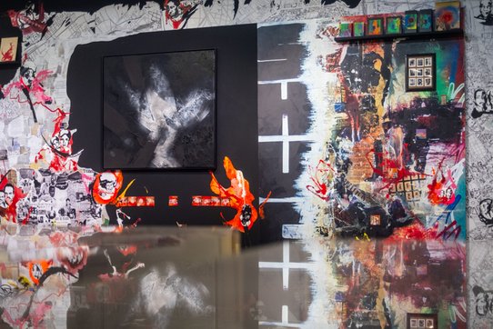 Cette fresque murale monumentale occupe tout un pan de mur, mêlant art urbain, abstraction et narration visuelle. À gauche, un fond composé de coupures de journaux en noir et blanc est envahi par des visages sérigraphiés expressifs, parfois entourés d'éclaboussures rouges et orangées. Au centre, une œuvre encadrée en noir, à dominante noire et grise, évoque une abstraction organique, semblable à des nuées ou des fractures rocheuses. À droite, une section de mur peinte présente une composition éclatée, avec des effets de feu et de ruine, des textures multiples, des silhouettes, des graffitis colorés, et une grille de petites images comme une mémoire fragmentée. Des touches de peinture fluorescente et des déchirures visuelles dynamisent l’ensemble. L’éclairage doux du lieu crée un effet de miroir dans les plexiglas d’autres œuvres exposées, amplifiant l’effet immersif de l’œuvre.