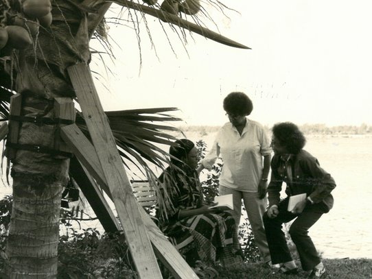 Beneath the wide shade of a coconut-laden palm tree, three women are engaged in a thoughtful and collaborative exchange. One of them, seated on a wooden bench, wears a traditional outfit with geometric patterns and holds an open notebook. The other two, dressed in casual shirts and trousers, stand or crouch nearby, attentively listening and holding their own papers. Their body language suggests a deep conversation—possibly an interview or oral testimony. In the background, a serene body of water outlines the edge of the scene, adding to the peaceful, outdoor atmosphere. The black-and-white composition enhances the timeless quality and historical depth of this encounter, likely centered on memory, heritage, and language, as part of Sarah Maldoror’s 1987 documentary dedicated to Aimé Césaire.