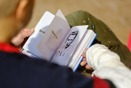 Shuck One, ici photographié par Pierre Malherbet, figure emblématique du street art, est ici photographié en train de consulter un carnet de croquis ouvert sur ses genoux. L’image, captée de dos, met en évidence ses mains tenant précautionneusement un carnet ligné dans lequel figure un dessin au trait noir : le portrait d’un homme au regard intense, coiffé avec élégance. Sur la page opposée, on distingue des inscriptions manuscrites telles que "Wifredo Lam" accompagnées de dates. Le fond neutre et flou concentre l’attention sur le carnet et les gestes de l’artiste, habillé d’un pull à rayures noires et rouges et d’un pantalon taché de peinture, signe de son immersion dans le processus créatif. Cette scène intime révèle une phase de réflexion ou de documentation dans le travail de Shuck One, mêlant mémoire, croquis préparatoire et hommage graphique.