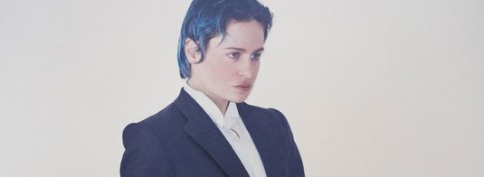 Mid-shot portrait of Rahim C. Redcar, also known as Christine and the Queens, wearing a structured black suit over a crisp white shirt. His intense, off-camera gaze and composed posture convey both theatrical presence and deep introspection. The slicked-back dark blue hair adds a bold, modern edge to his androgynous silhouette. The plain, light background enhances the minimalist elegance and draws focus to the unique identity and artistic style of the performer, blending classic formality with personal expression.