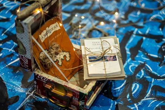 Au centre de cette installation artistique, une boîte réalisée à partir de canettes de soda recyclées accueille deux livres anciens soigneusement attachés avec de la ficelle naturelle. L’un, de couleur orange usée, est intitulé « La Martinique, Grand Océan », et présente une silhouette de l’île en relief entourée d’un fil de jute. L’autre, de tons beiges et blancs, porte le titre « Pays rêvé, pays réel » de l’écrivain Édouard Glissant, dont une photo partielle orne la couverture. La boîte repose sur un sol couvert de motifs calligraphiques noirs sur fond bleu indigo, emblématique du travail de Valérie John, évoquant la teinture naturelle utilisée historiquement en Afrique et dans les Antilles. De fines guirlandes lumineuses serpentent autour des objets, ajoutant une dimension onirique et sacrée à la scène. Cette composition, à la fois poétique et politique, fait partie de l’œuvre-installation multimédia SECRET(S)... RÊVES DE PAYS... FABRIQUE À MÉMOIRE(S)... PALIMPSESTE... (1998-2025), une commande du Centre Pompidou qui explore les liens entre mémoire, territoire, identité postcoloniale et héritages culturels.