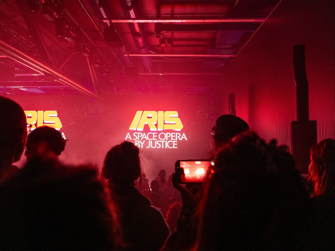 Dans une salle obscure aux teintes rouges profondes, le public découvre le titre "IRIS – A Space Opera by Justice" projeté sur un écran monumental. L’ambiance est futuriste, immersive, presque cinématographique. La fumée diffuse accentue la sensation d’apesanteur, et les silhouettes capturent la scène via leurs smartphones, suspendues dans l’attente d’un voyage audiovisuel signé par le duo électro Justice.