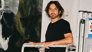 L’artiste Lionel Sabatté se tient sur une nacelle élévatrice dans son atelier, devant une immense toile abstraite aux tonalités sombres et dorées. À ses côtés, des échelles et câbles témoignent du travail en cours d’accrochage ou de création monumentale.