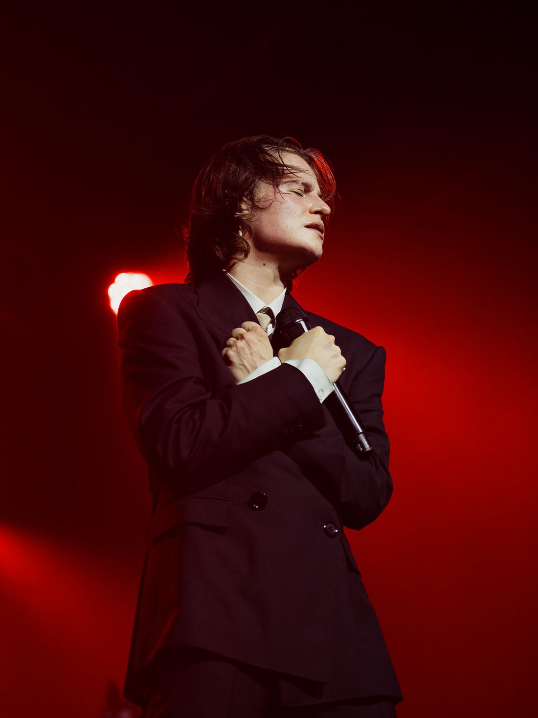 Christine and the Queens, en costume noir sobre, incarne l’intensité émotionnelle sur scène pendant "Because Beaubourg". Les bras croisés sur le cœur, les traits marqués par la lumière rouge, l’artiste semble suspendu dans une performance où chaque geste devient expression. Cette posture théâtrale et poignante, sublimée par l’éclairage dramatique, capture toute la puissance scénique et poétique du personnage.