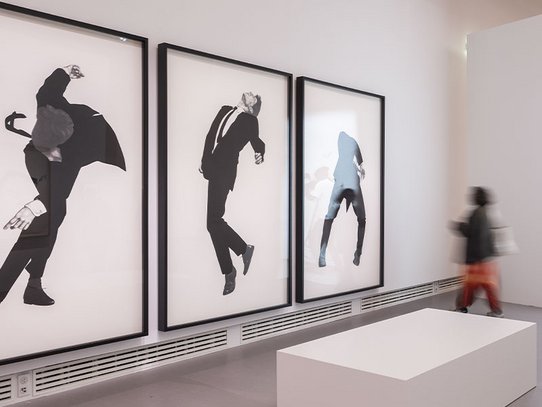 Inside a minimalist room at the Grand Palais, three large black-and-white drawings by Robert Longo portray suited men frozen in explosive, theatrical poses. The suspended motion conveys intense emotion and dynamic tension. A blurred visitor walks past, injecting fleeting movement into the otherwise static gallery, amplifying the visual power of this impactful triptych.