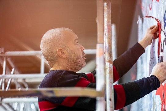 L’artiste urbain Shuck One, pris en photo par Pierre Malherbet, en pleine action, concentré sur la réalisation d’une fresque murale. Il se tient debout sur un échafaudage, les bras levés, appliquant soigneusement un élément graphique sur le mur. Son regard est fixe, déterminé, révélant une implication totale dans son geste artistique. Vêtu d’un pull à larges bandes noires et bordeaux, il se détache légèrement de l’arrière-plan flou mais coloré, où l’on devine des éclaboussures de peinture et des formes abstraites. À ses côtés, la structure métallique de l’échafaudage évoque l’aspect technique et physique de l’œuvre murale. La lumière ambiante, douce et légèrement orangée, accentue l’atmosphère de création intense et silencieuse, comme suspendue dans le temps. Cette scène illustre l’engagement manuel et émotionnel de l’artiste dans son processus de création.