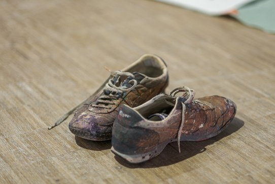 Posées négligemment sur un sol en bois, cette paire de chaussures porte les marques visibles du temps et de la création. Couvertes d’éclaboussures de peinture multicolore — des teintes de violet, rouge, ocre et noir — les baskets usées témoignent du quotidien de l’artiste Shuck One. Chaque tache, chaque trace semble raconter une partie de son processus artistique, comme une extension silencieuse de ses gestes sur la toile ou le mur. La semelle usée, les lacets défaits et la texture du cuir vieilli renforcent cette impression d’authenticité brute, de vécu. Cette image minimaliste mais parlante incarne la matérialité de l’art urbain : un travail au sol, en hauteur, en mouvement, dans lequel l’artiste s’immerge pleinement. Plus qu’un simple accessoire, ces chaussures deviennent une empreinte discrète mais puissante de la démarche créative de Shuck One.