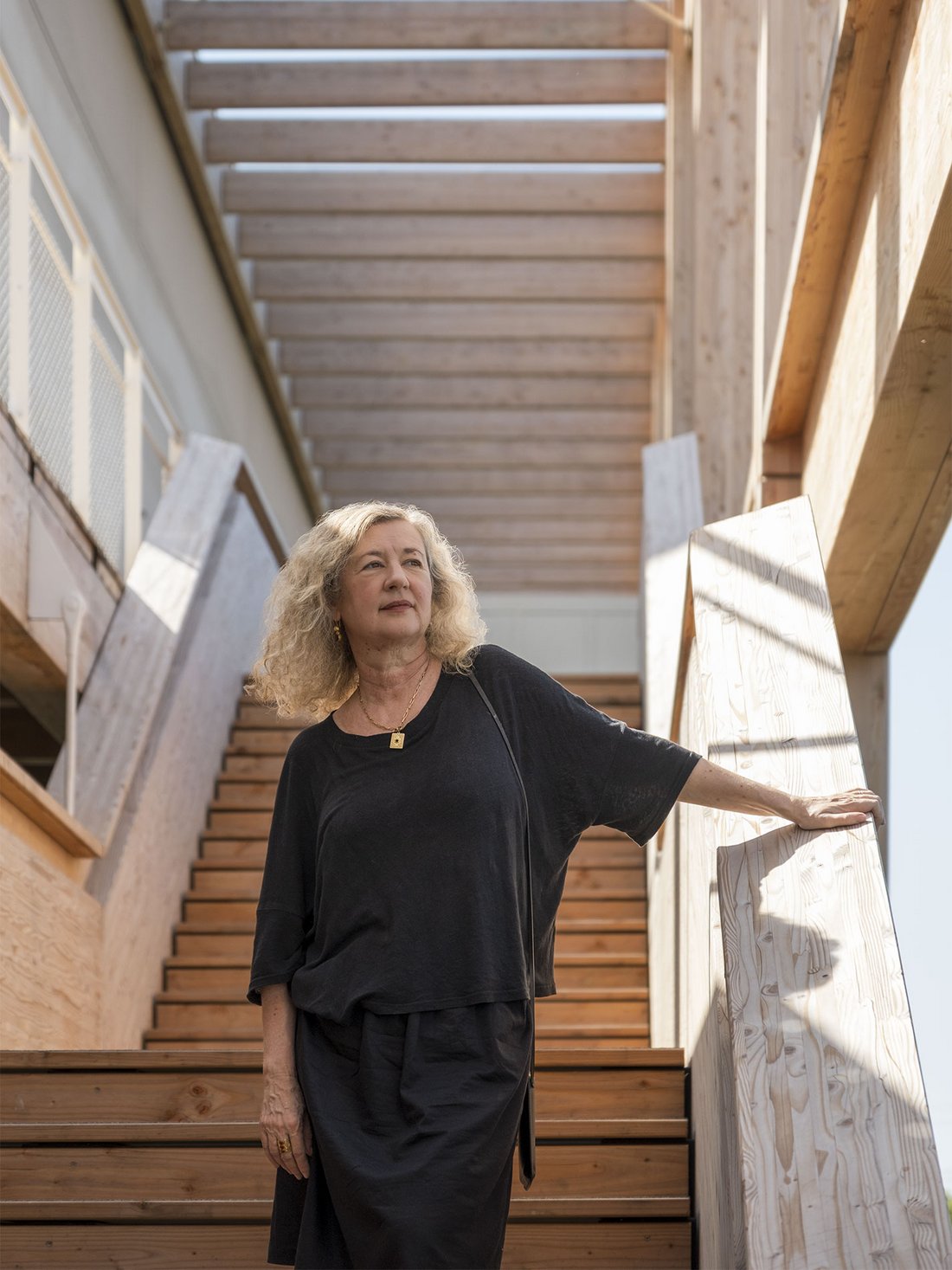 Portrait d’Alexia Fabre, directrice du Centre Pompidou Francilien, debout dans un escalier monumental en bois. Vêtue de noir, elle s’appuie sur une rambarde et regarde sur le côté, dans une architecture baignée de lumière naturelle. Photo : Pierre Malherbet.
