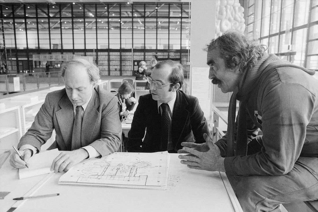 Cette photographie en noir et blanc, capturée dans le cadre du montage de l’exposition « Le Crocrodrome de Zig et Puce », montre Jean Tinguely en pleine discussion avec deux collaborateurs, très probablement architectes ou commissaires d’exposition. Tinguely, reconnaissable à sa moustache fournie, ses cheveux en bataille et sa tenue décontractée (pull et écharpe), est penché en avant, les mains expressives, commentant avec intensité les plans étalés sur la table. Face à lui, deux hommes en costume-cravate l’écoutent attentivement : l’un prend des notes, l’autre observe le plan avec concentration. Le dessin visible sur la table semble être une vue d’implantation de l’installation dans l’espace du Centre Pompidou, mêlant schéma technique et indications manuscrites. En arrière-plan, les volumes ouverts et modulaires de la bibliothèque publique du centre (BPI) témoignent de l’architecture innovante du bâtiment. Cette scène capture à la fois l’aspect collaboratif du projet, la rigueur de la préparation technique, et l’énergie charismatique de l’artiste engagé dans l’une de ses œuvres les plus spectaculaires.
