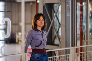 Émilie Tronche, dessinatrice connue notamment pour sa série « Samuel », pose avec assurance dans un espace emblématique du Centre Pompidou. Vêtue d’un haut de survêtement violet chiné aux détails bordeaux et d’un jean taille haute, elle s’appuie légèrement contre une rambarde blanche en métal ajouré. Son regard franc et tranquille est dirigé vers l’objectif, tandis que la lumière ambiante met en valeur ses traits et son style sobre et contemporain. En arrière-plan, les structures métalliques croisées et les parois vitrées caractéristiques du bâtiment dialoguent avec une grande lettre « C » suspendue et lumineuse, renforçant l’ambiance artistique et architecturale du lieu. Cette photo, prise par Pierre Malherbet, capte un instant d’intimité dans un cadre emblématique de la scène culturelle parisienne.