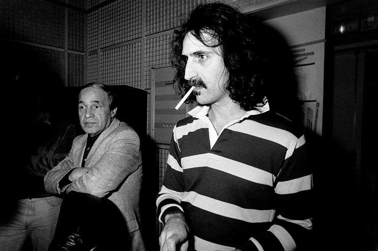 Dans une pièce tapissée de panneaux acoustiques, deux légendes de la musique contemporaine se tiennent côte à côte, incarnant deux mondes artistiques qui se rencontrent. À droite, Frank Zappa, cheveux longs et moustache caractéristique, porte un polo à larges rayures noires et blanches. Il tient une cigarette entre les lèvres, les mains engagées dans un geste suggérant la préparation ou l’attention technique. Son regard est fixé, concentré, absorbé dans l’instant. À gauche, Pierre Boulez, figure austère et respectée de la musique contemporaine française, croise les bras et observe calmement la scène. Vêtu d’une veste claire, son expression est à la fois distante et attentive, illustrant son rôle de chef d’orchestre et de penseur musical. L’image en noir et blanc renforce la gravité du moment, documentant une collaboration rare entre l’avant-garde américaine et l’avant-garde européenne, probablement lors de l'enregistrement de l'album The Perfect Stranger en 1984, dirigé par Boulez et composé par Zappa.