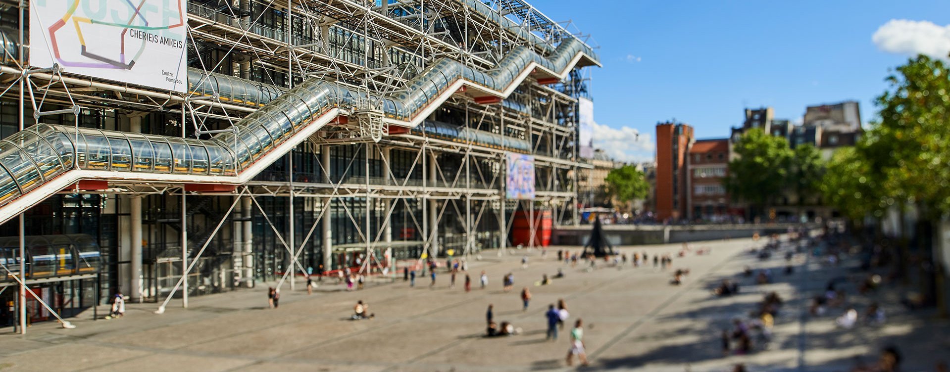 The Centre Pompidou in Paris under a bright blue sky, showcasing its iconic metal structure and transparent tubular escalators overlooking a lively square, The Piazza, filled with pedestrians and tourists.