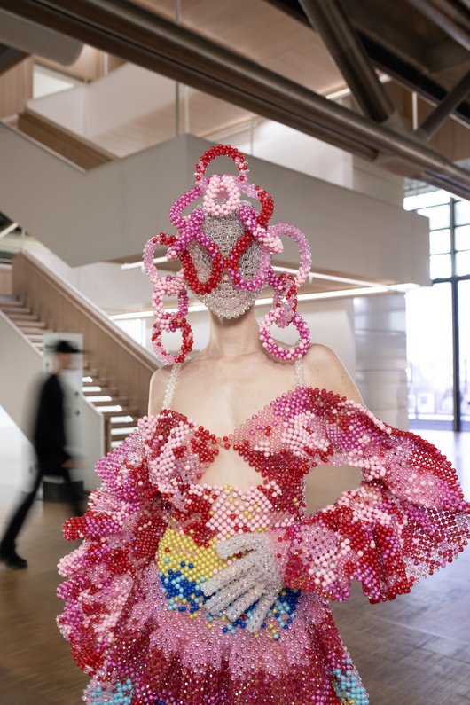 Exposition "La traversée des apparences. La mode au Musée" – affiche. Mannequin portant une robe et un masque ornés de perles roses et rouges, exposé dans le Centre Pompidou