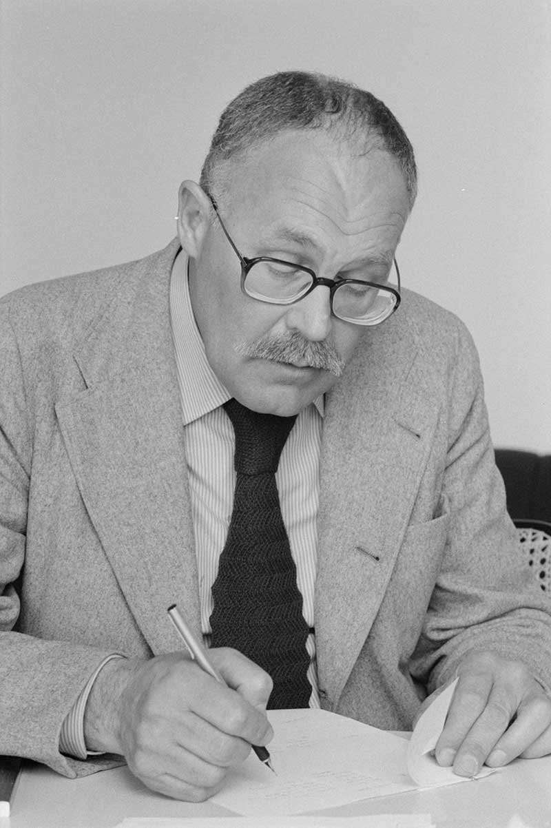 This black-and-white photograph captures Pontus Hulten in June 1977 at the Centre Georges Pompidou in Paris. The Swedish art historian and curator is seen seated at a desk, deeply focused as he writes on several sheets of paper in front of him. Dressed in a grey wool suit, a striped shirt, and a dark knitted tie, Hulten embodies the blend of elegance and intellectual discipline. His round glasses sit low on his nose as he leans forward, immersed in his work. His strong hands hold a fountain pen, and one corner of the paper is folded beneath his left hand. The minimalist setting reflects the sober administrative environment of a newly established cultural institution. Captured only a few months after the Centre Pompidou’s public opening, the image conveys the foundational role Hulten played as its first director — a pioneer navigating the intersection of contemporary art, cultural politics, and visionary museology.