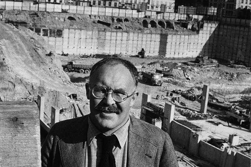 Pontus Hulten, a pivotal figure in the contemporary art world, stands in the foreground of the bustling construction site of the Centre Pompidou in Paris, circa 1975. Dressed in a wool suit, light shirt, and loosely tied black necktie, he gazes confidently into the camera with a subtle, knowing smile. His thick mustache and round glasses enhance his intellectual and determined presence. Behind him, the vast excavated site reveals the foundational phase of what would become one of the most radical cultural institutions of the 20th century. Dump trucks, cranes, and rising concrete elements animate the pit, framed by towering retaining walls. Above the site, classic Haussmann-style buildings underline the stark contrast between tradition and architectural modernity. Captured in black and white, the image encapsulates a historical moment of transformation, with Hulten symbolizing the merger of artistic vision and urban innovation as the founding director of the Centre Pompidou.