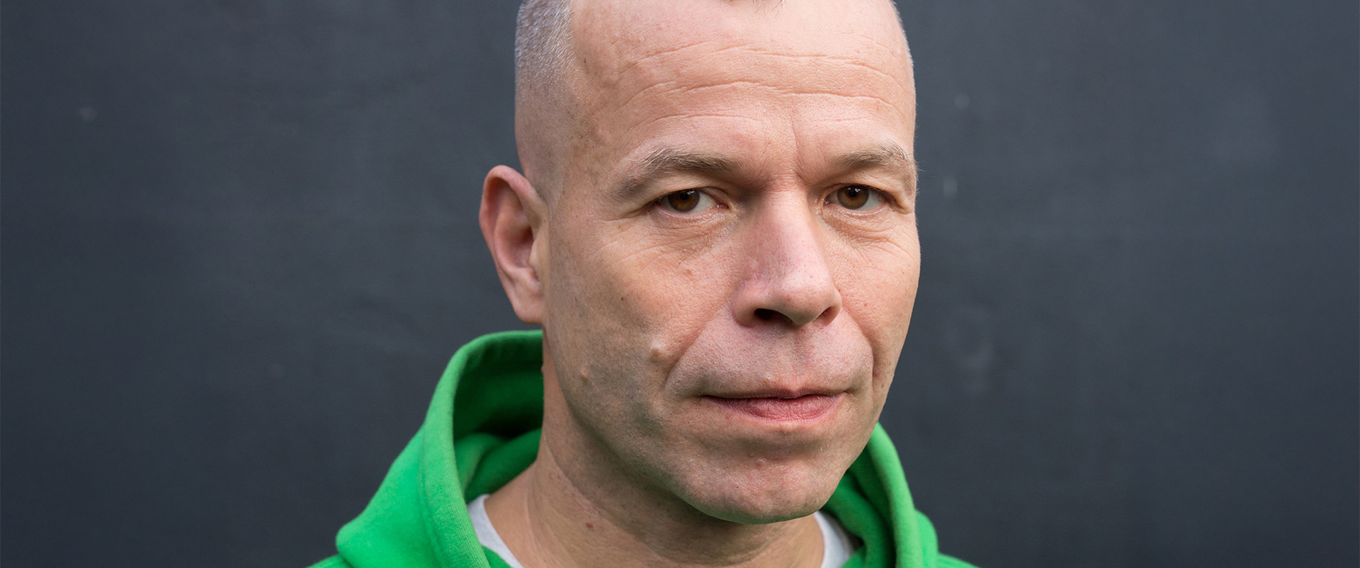 Ce portrait de Wolfgang Tillmans présente l'artiste dans une posture frontale, regardant droit vers l’objectif avec une expression calme et légèrement sérieuse. Il porte un sweat-shirt à capuche vert vif, contrastant vivement avec le fond neutre gris foncé. La lumière naturelle et douce met en valeur les traits du visage de Tillmans : ses yeux clairs, ses rides d’expression discrètes, et la texture naturelle de sa peau, témoignage d'une approche sincère et sans artifices. L’arrière-plan sombre et uni renforce la présence immédiate du sujet, recentrant toute l'attention sur son visage et son attitude posée. Ce type de portrait minimaliste, direct et non théâtral est en parfaite cohérence avec l’éthique visuelle de Tillmans, souvent axée sur l'authenticité, la spontanéité et la transparence. L’atmosphère générale évoque la simplicité et la profondeur du regard d'un artiste ayant profondément transformé le langage photographique contemporain.