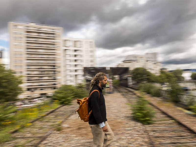 Alain Gomis marche sur les rails abandonnés de la Petite Ceinture, dans le 19e arrondissement de Paris. Sac en cuir sur l’épaule, il traverse un paysage urbain flou, entre immeubles modernes, végétation sauvage et ciel chargé de nuages. L’image de Pierre Malherbet évoque le mouvement, le voyage, la mémoire et une atmosphère cinématographique.