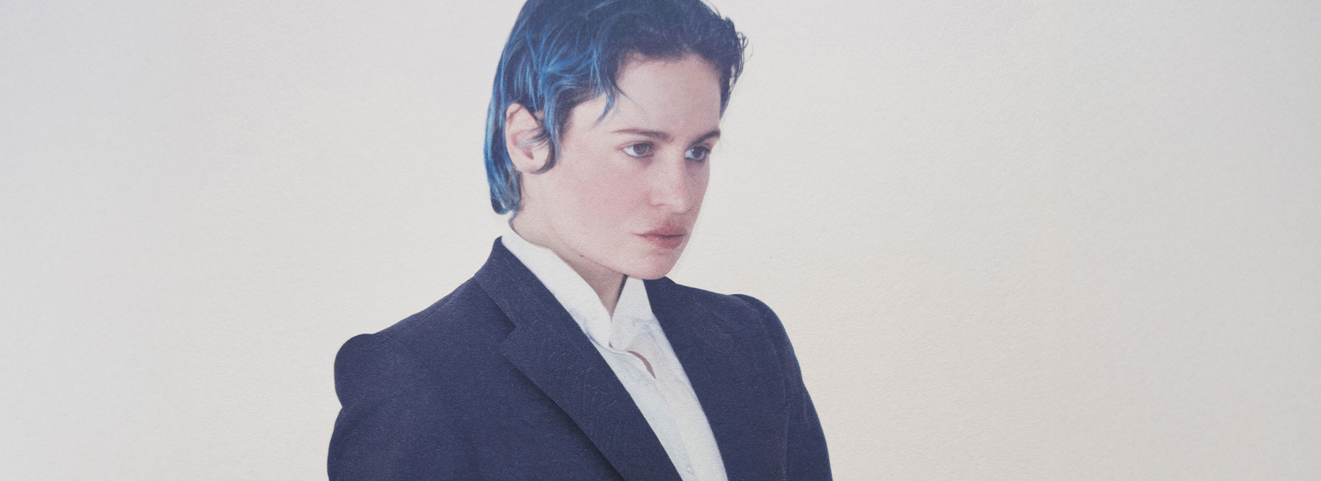 Portrait en plan moyen de Rahim C. Redcar, alias Christine and the Queens, vêtu d’un costume noir structuré sur une chemise blanche immaculée. Son regard intense, dirigé hors champ, et sa posture soignée traduisent une présence à la fois théâtrale et introspective. Ses cheveux bleu foncé, plaqués vers l’arrière, apportent une touche moderne et audacieuse à cette silhouette androgyne. Le fond uni, clair, renforce la sobriété de la composition et met en lumière l’esthétique singulière de l’artiste, entre élégance classique et affirmation identitaire.