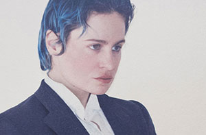 Portrait en plan moyen de Rahim C. Redcar, alias Christine and the Queens, vêtu d’un costume noir structuré sur une chemise blanche immaculée. Son regard intense, dirigé hors champ, et sa posture soignée traduisent une présence à la fois théâtrale et introspective. Ses cheveux bleu foncé, plaqués vers l’arrière, apportent une touche moderne et audacieuse à cette silhouette androgyne. Le fond uni, clair, renforce la sobriété de la composition et met en lumière l’esthétique singulière de l’artiste, entre élégance classique et affirmation identitaire.