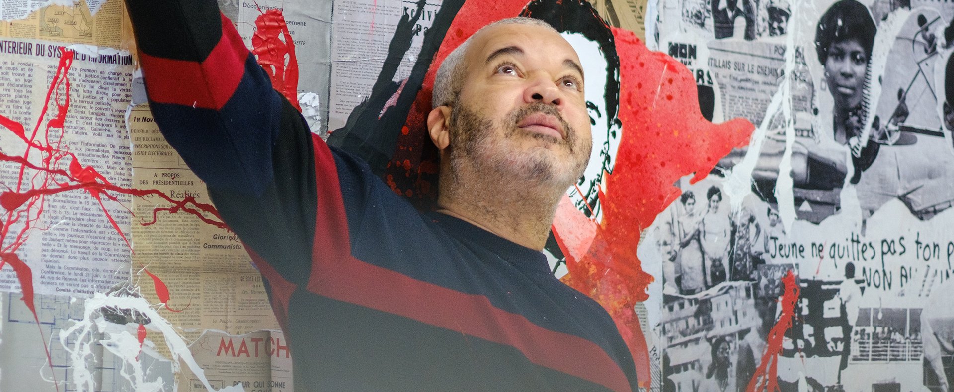 L’artiste Shuck One, photographié par Pierre Malherbet, se tient debout devant une fresque dense et expressive, composée d’un collage de coupures de presse historiques, d’illustrations en noir et blanc, et de projections de peinture rouge et noire. Le mur derrière lui évoque à la fois la mémoire collective, les luttes sociales et l’expression urbaine, mêlant des unes de journaux anciens, des visages emblématiques, et des slogans évocateurs comme "Jeune, ne quittes pas ton pays, non à !". Vêtu d’un pull bleu marine à bandes bordeaux, Shuck One pointe vers le haut d’un geste vif, le regard tourné dans la même direction, comme pour souligner ou questionner un élément précis de l’œuvre. L’ambiance est intense, à la croisée de l’art urbain et de l’histoire, avec une énergie revendicative palpable à travers les éclaboussures rouges qui sillonnent la composition.