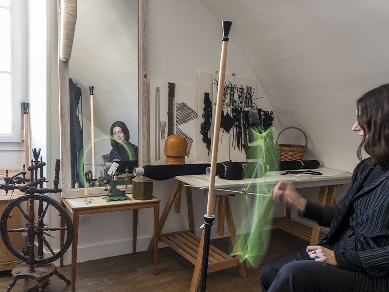 Cette photographie plonge dans l’univers de création de Jeanne Vicérial au cœur de son atelier textile. L’artiste est assise dans un espace baigné de lumière naturelle, entourée de métiers artisanaux, de bobines de fil, de structures textiles et d’échantillons noirs accrochés au mur. Son geste en mouvement crée une trace lumineuse verte qui traverse l’image comme une énergie vivante ou un dessin immatériel. Le miroir placé à gauche reflète discrètement son visage concentré, renforçant l’idée de double regard entre création et introspection. Les matières textiles, les outils anciens et les lignes épurées de l’atelier composent une atmosphère à la fois expérimentale et intime. L’image traduit le dialogue entre artisanat traditionnel, innovation textile et recherche artistique contemporaine, dans une esthétique calme, sensible et profondément humaine.