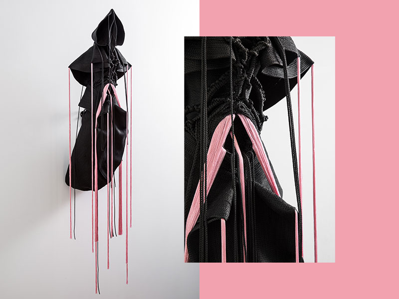 L’œuvre textile Mue de Jeanne Vicérial se présente comme une sculpture suspendue mêlant artisanat, mode expérimentale et recherche contemporaine sur le textile. Réalisée dans des tons noirs profonds contrastés par de fines bandes roses, la pièce déploie des volumes organiques, des plis sculpturaux et de longues franges verticales qui accentuent l’impression de mouvement et de transformation. Les matières tressées et nouées évoquent à la fois une armure souple, une silhouette vivante et une structure en mutation permanente. Le fond minimaliste met en valeur la sophistication des textures et la précision du travail manuel. Le détail rapproché révèle la richesse des fibres et des entrelacements, renforçant l’aspect tactile et sensoriel de l’œuvre. Entre couture, sculpture et design textile, Mue incarne une réflexion artistique autour du corps, de la métamorphose et de la mémoire des matières.