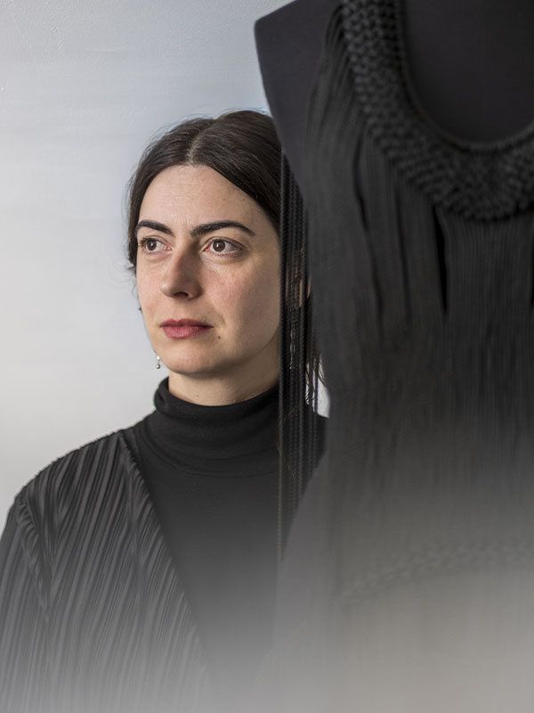 Portrait intimiste de l’artiste et designer textile Jeanne Vicérial, photographiée dans une lumière douce et minimaliste par Pierre Malherbet. Son regard calme et concentré se dirige hors champ, tandis qu’une œuvre noire aux textures tressées et frangées occupe le premier plan. Les contrastes entre la peau claire, les vêtements noirs plissés et les matières textiles créent une atmosphère élégante, contemporaine et silencieuse. L’image évoque la création artisanale, la mode expérimentale et le dialogue entre corps, couture et sculpture. Le cadrage épuré met en valeur la sensibilité artistique de l’artiste ainsi que la sophistication des matériaux et du travail textile.
