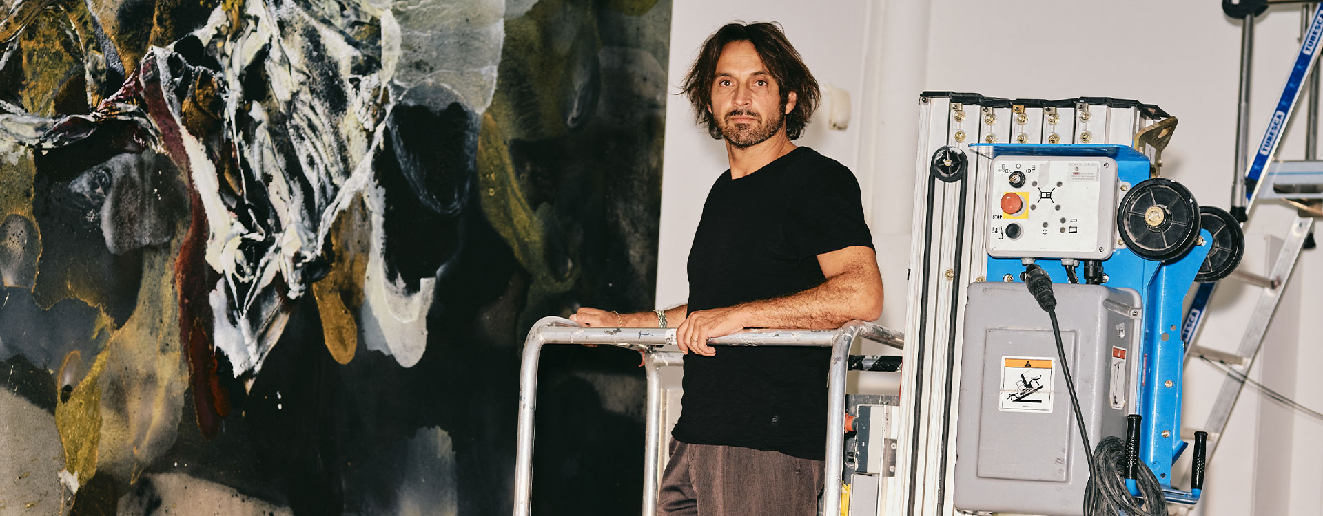 L’artiste Lionel Sabatté se tient sur une nacelle élévatrice dans son atelier, devant une immense toile abstraite aux tonalités sombres et dorées. À ses côtés, des échelles et câbles témoignent du travail en cours d’accrochage ou de création monumentale.