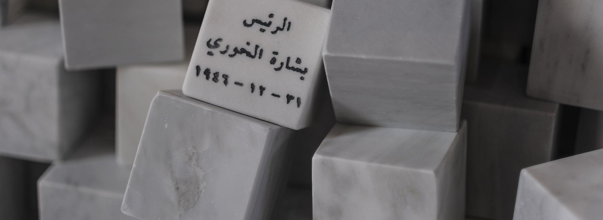 Dans une salle d’exposition baignée d’ombre, un imposant amas de cubes en marbre blanc se dresse en forme de pyramide irrégulière. L’œuvre, signée par l’artiste Marwan Rechmaoui, est exposée à Auxerre dans le cadre de l’exposition "Quête d’infinis". Chaque cube, aux arêtes nettes et aux veines subtiles typiques du marbre, semble représenter une entité historique ou une figure de pouvoir : on y lit, gravés avec sobriété, des noms comme "Sultan Abdul Hamid II 1876–1909", "President Alfred Agatecha 22-12-1963" ou encore "The Allied Forces 1941". Certains sont inscrits en arabe, renforçant la portée géopolitique et mémorielle de l’installation. Les blocs forment un tumulus ordonné mais fragmenté, où les éléments tombés ou décentrés suggèrent les ruines, les couches de l’histoire ou l’instabilité du pouvoir. Une lumière naturelle filtre par une fenêtre en bois, projetant sur le sol des ombres graphiques qui contrastent avec la froide pureté minérale des cubes. L’ensemble évoque un monument silencieux à la mémoire des empires, des nations et des figures marquantes du monde arabe et occidental.