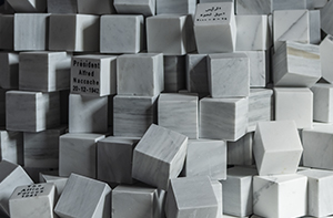Dans une salle d’exposition baignée d’ombre, un imposant amas de cubes en marbre blanc se dresse en forme de pyramide irrégulière. L’œuvre, signée par l’artiste Marwan Rechmaoui, est exposée à Auxerre dans le cadre de l’exposition "Quête d’infinis". Chaque cube, aux arêtes nettes et aux veines subtiles typiques du marbre, semble représenter une entité historique ou une figure de pouvoir : on y lit, gravés avec sobriété, des noms comme "Sultan Abdul Hamid II 1876–1909", "President Alfred Agatecha 22-12-1963" ou encore "The Allied Forces 1941". Certains sont inscrits en arabe, renforçant la portée géopolitique et mémorielle de l’installation. Les blocs forment un tumulus ordonné mais fragmenté, où les éléments tombés ou décentrés suggèrent les ruines, les couches de l’histoire ou l’instabilité du pouvoir. Une lumière naturelle filtre par une fenêtre en bois, projetant sur le sol des ombres graphiques qui contrastent avec la froide pureté minérale des cubes. L’ensemble évoque un monument silencieux à la mémoire des empires, des nations et des figures marquantes du monde arabe et occidental.