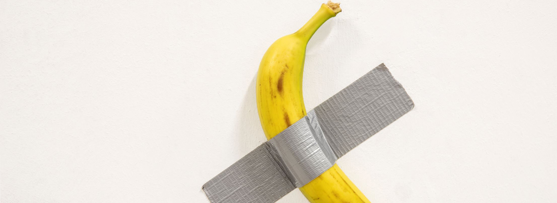 Une banane jaune tachetée de brun, bien mûre, est scotchée de manière brute contre un mur blanc uni. Elle est maintenue par un large morceau de ruban adhésif gris disposé en biais, créant une croix imparfaite. L'œuvre joue volontairement sur la simplicité et l’absurde, évoquant une esthétique minimaliste et provocante. L’absence d’arrière-plan ou d’élément décoratif met en valeur la nature décalée de l’objet, emblématique de l’art contemporain conceptuel. Cette image fait directement référence à l’œuvre « Comedian » de Maurizio Cattelan, célèbre pour sa critique de la marchandisation de l’art et sa capacité à susciter débat et réflexion à partir d’un objet banal.