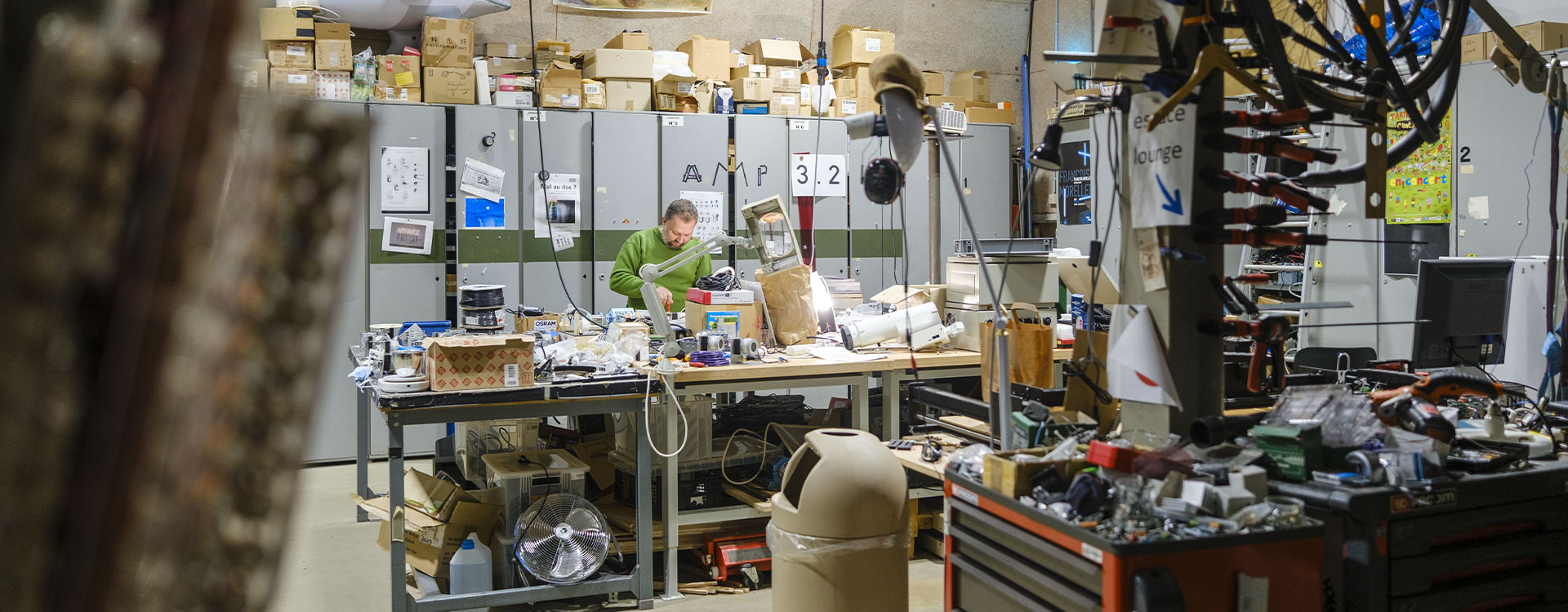 L’image montre un vaste atelier foisonnant d’énergie créative, où un artisan ou technicien en pull vert se penche sur un plan de travail encombré d’outils, de câbles et de composants électroniques. Les étagères et les armoires métalliques à l’arrière débordent de cartons et de matériaux, témoignant d’une activité intense et expérimentale. Au premier plan, des roues de vélo, des pinces et des boîtes d’outils soulignent la dimension manuelle et technique du lieu. Les affiches artistiques accrochées au mur ajoutent une touche culturelle, révélant un espace hybride entre art, invention et artisanat industriel.