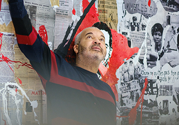 L’artiste Shuck One se tient debout devant une fresque dense et expressive, composée d’un collage de coupures de presse historiques, d’illustrations en noir et blanc, et de projections de peinture rouge et noire. Le mur derrière lui évoque à la fois la mémoire collective, les luttes sociales et l’expression urbaine, mêlant des unes de journaux anciens, des visages emblématiques, et des slogans évocateurs comme "Jeune, ne quittes pas ton pays, non à !". Vêtu d’un pull bleu marine à bandes bordeaux, Shuck One pointe vers le haut d’un geste vif, le regard tourné dans la même direction, comme pour souligner ou questionner un élément précis de l’œuvre. L’ambiance est intense, à la croisée de l’art urbain et de l’histoire, avec une énergie revendicative palpable à travers les éclaboussures rouges qui sillonnent la composition.