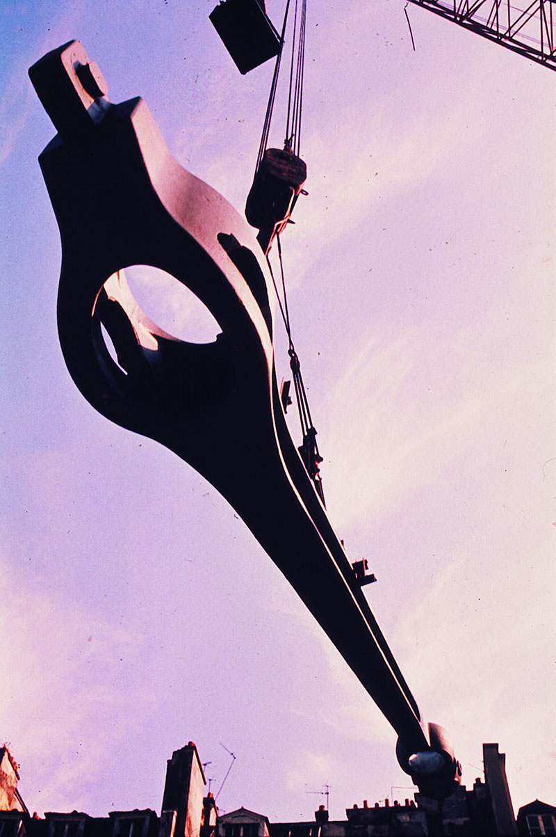 Dans une lumière crépusculaire teintée de rose, une gerberette monumentale en acier est en cours de levage par une grue, suspendue au-dessus des toits parisiens. Cette pièce emblématique du Centre Pompidou illustre l’audace architecturale du projet. Le contraste entre la masse imposante de la structure et la douceur du ciel accentue l’esthétique industrielle du mouvement high-tech. La scène capture un instant suspendu, à la croisée de l’ingénierie et de l’art.