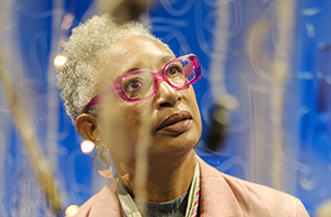 Valérie John, artiste plasticienne et créatrice engagée, est ici photographiée en pleine immersion dans son œuvre, présentée dans le cadre de l’exposition « Paris noir » au Centre Pompidou. Vêtue d’une veste rose pâle et d’une cravate graphique décorée de pendentifs symboliques — un cœur incrusté et une figure dorée — elle se tient droite, les mains derrière le dos, le regard tourné vers le haut dans une posture de contemplation intérieure. Ses lunettes roses translucides, assorties à sa tenue, ajoutent une touche vive à son expression concentrée. Autour d’elle, des fils suspendus portent des objets organiques ou artisanaux — galets, coquillages, pièces sculptées, perles — dans une installation aérienne, comme une constellation intime flottant dans un espace bleu profond. Les lumières chaudes et tamisées reflétées sur certains éléments créent une ambiance onirique, entre spiritualité, mémoire et ancrage culturel. L’image traduit à la fois la puissance de la présence de l’artiste dans son œuvre et la richesse sensorielle d’une installation qui convoque les récits diasporiques, l’héritage caribéen et la poésie des matières.