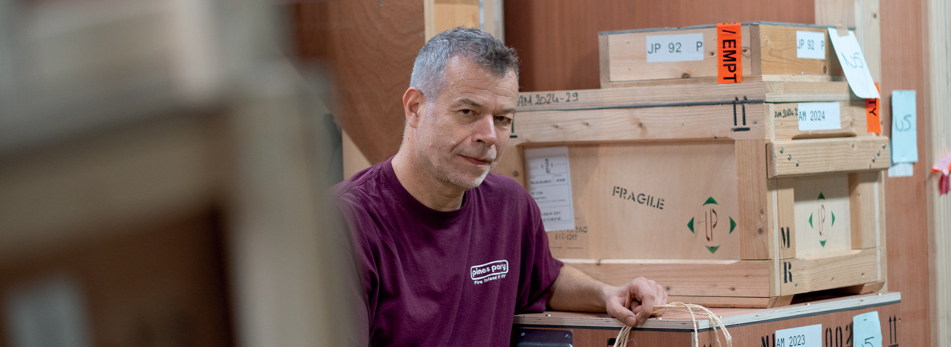 La scène capturée par Pierre Malherbet montre Wolfgang Tillmans dans les réserves du Centre Pompidou, entouré de grandes caisses en bois servant au transport sécurisé d’œuvres d’art. Tillmans, vêtu d’un t-shirt bordeaux arborant l'inscription 'Démé à Paris', se tient avec décontraction contre une caisse marquée 'Fragile'. Dans sa main gauche, il tient une bobine de cordelette d’emballage, symbole discret des préparatifs logistiques liés à l'installation de son exposition. L'expression sur son visage, à la fois concentrée et légèrement malicieuse, trahit l’intimité et la spontanéité de ce moment en coulisses. À l’arrière-plan, les empilements de caisses étiquetées évoquent l’effervescence invisible qui précède l'ouverture d'une grande exposition. Les tons chauds du bois brut et la lumière diffuse instaurent une atmosphère chaleureuse, presque artisanale, contrastant avec la rigueur froide souvent associée aux espaces de stockage muséaux.